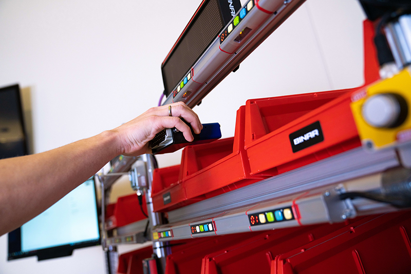 A person's hand is using a pick2light system, placing a object into a red bin that is part of an array of bins with digital displays indicating stock levels or item numbers.