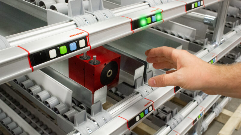 A hand points to a 'Pick to Light' system in a warehouse, with illuminated green and red light indicators, showcasing an efficient order picking technology that aids in quick and accurate item selection.
