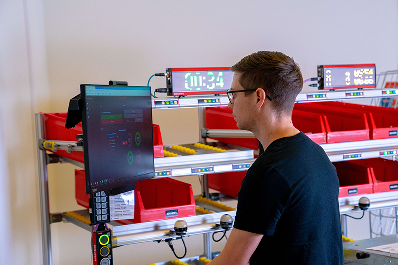 A focused worker is monitoring a pick2light system through a computer terminal, with a backdrop of organized red bins and electronic displays showing inventory status or order information.